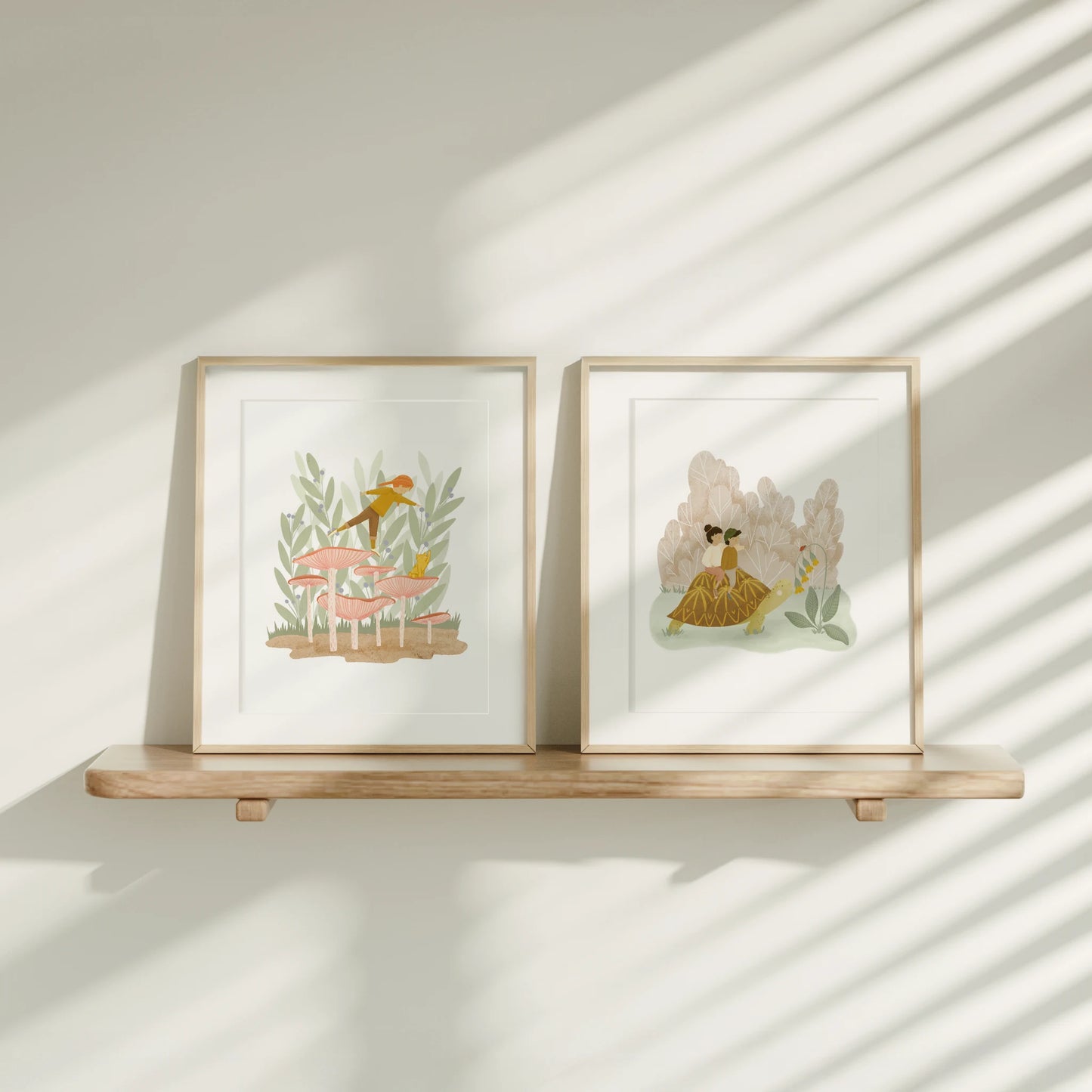 two art prints sitting on a wooden ledge in the sunlight, shown framed. on the left is 'leaning in' girl with mushroom and on the right is 'in no hurry' with 2 people riding a turtle.