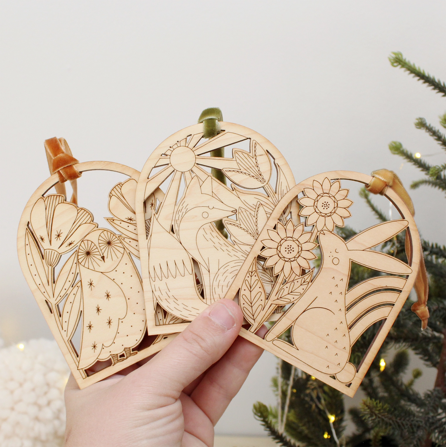 collection of wooden arch shaped insect ornaments with intricately etched designs: from left to right a owl, fox and rabbit, being held by a hand in front of a christmas tree.