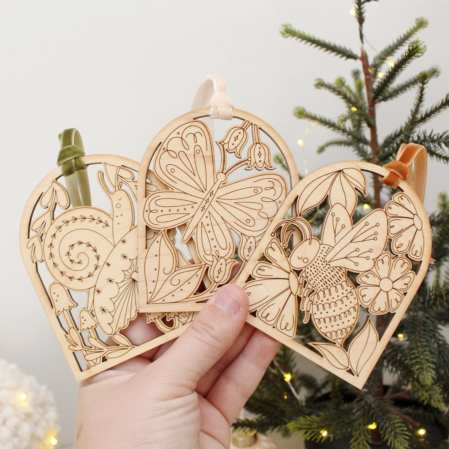 collection of wooden arch shaped insect ornaments with intricately etched designs: from left to right a snail, butterfly and bee, being held by a hand in front of a christmas tree.