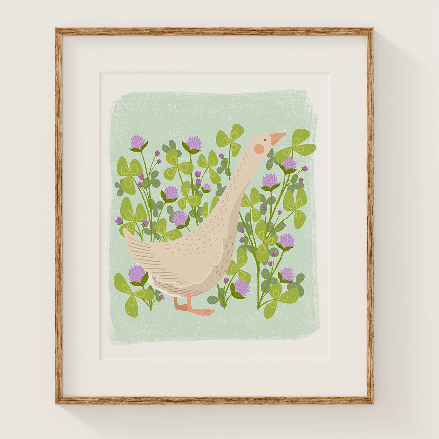 'meadow muse' art print, shown in a wood frame. print features a whimsical goose in front of a clovers and clover flowers.
