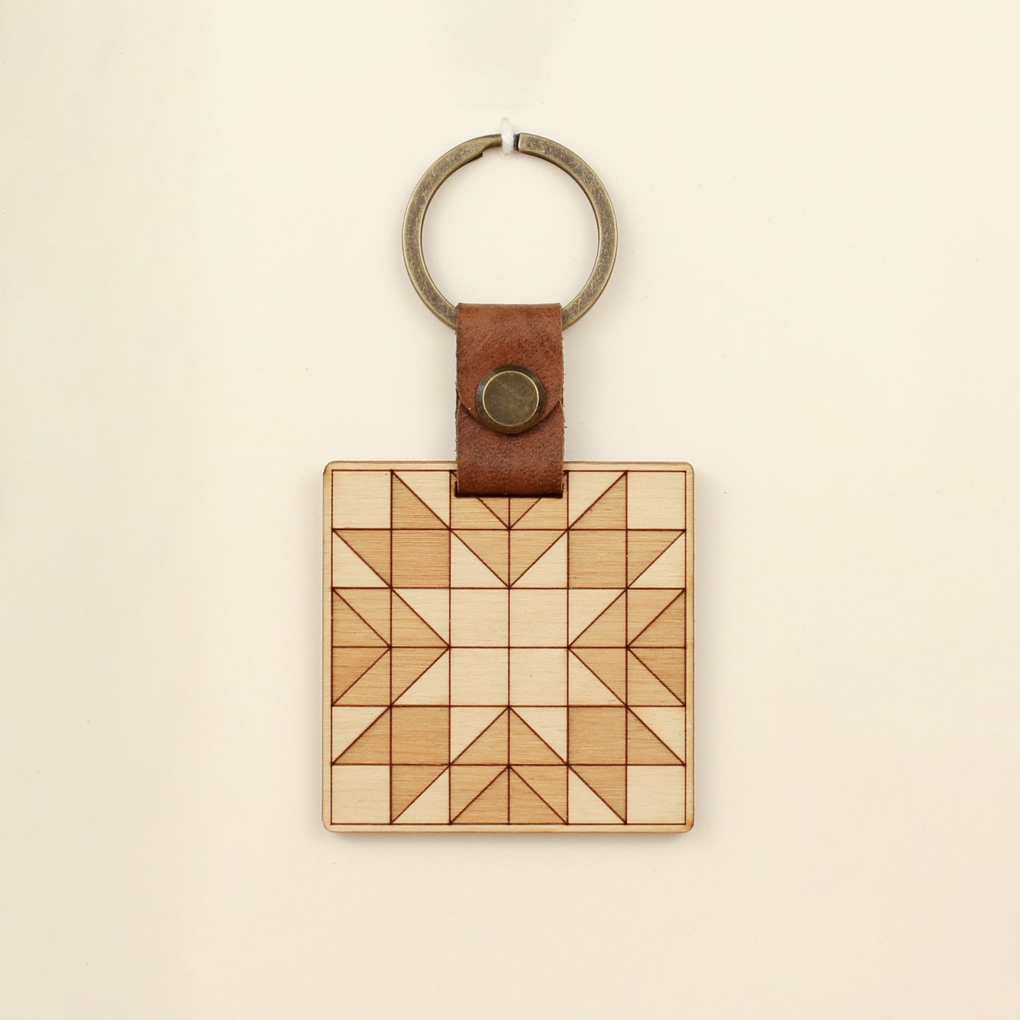 wooden keychain with quilt block pattern in two wood tones, with leather strap and bronze colored details.