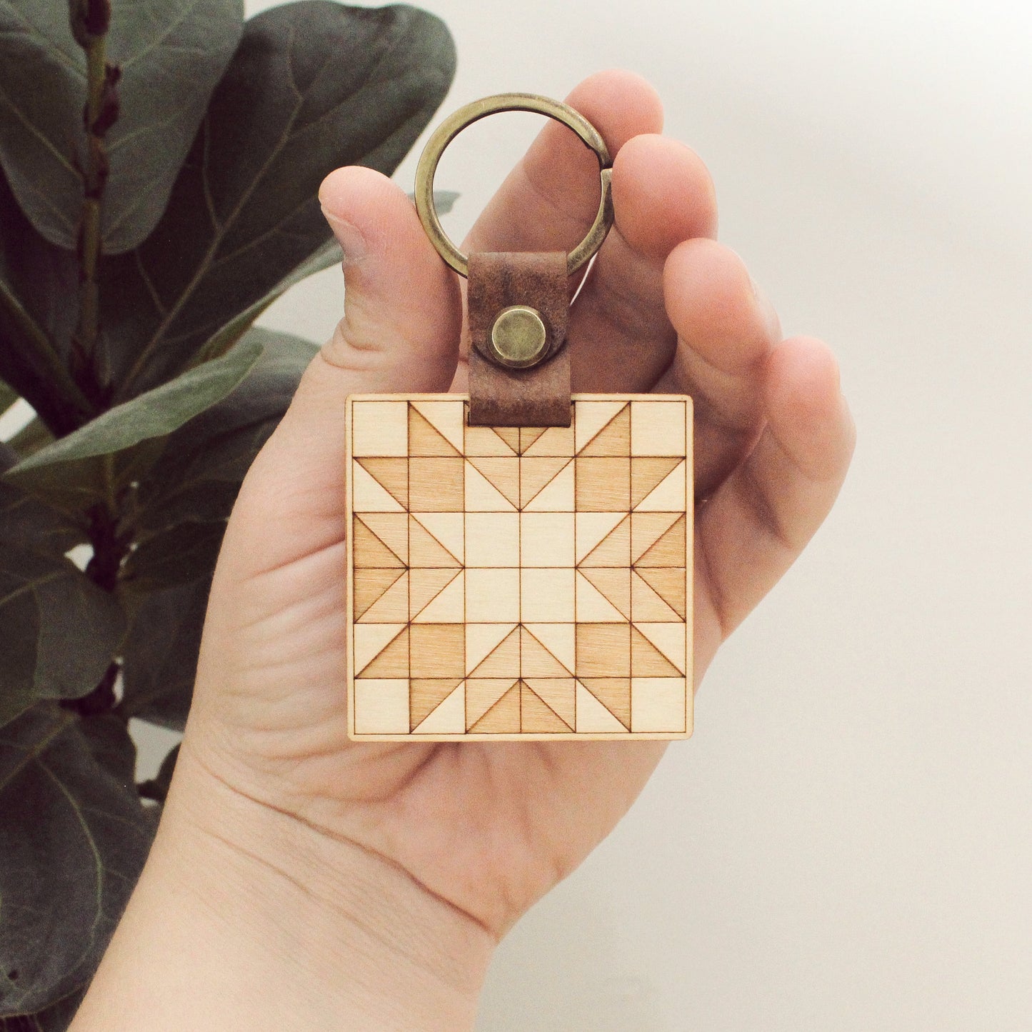 wooden keychain with quilt block pattern in two wood tones, with leather strap and bronze colored details.