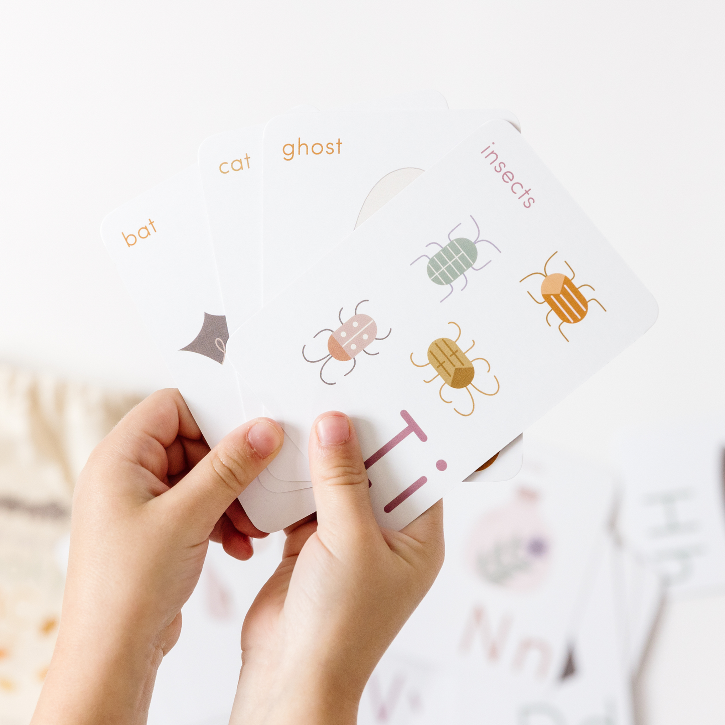 child's hands hold halloween ABC flashcards fanned out, from left to right are B bat, C cat, G ghost, and I insects.