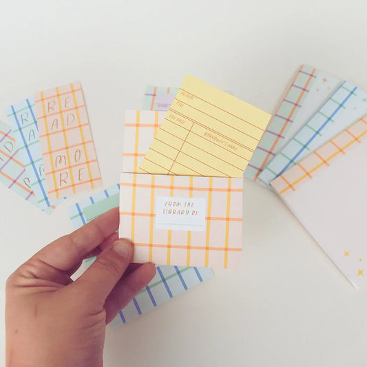 a hand holds a printed library book pocket. in the background are bookmarks and mini notepads. all designs are colorful rainbow plaids.