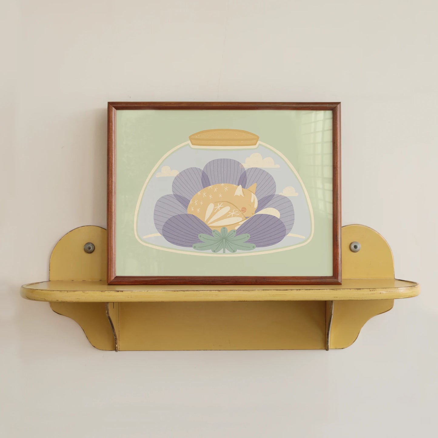 'a nap or two' art print, shown in a wood frame sitting on a yellow shelf. print features cat sleeping inside of a flower, inside of a jar.