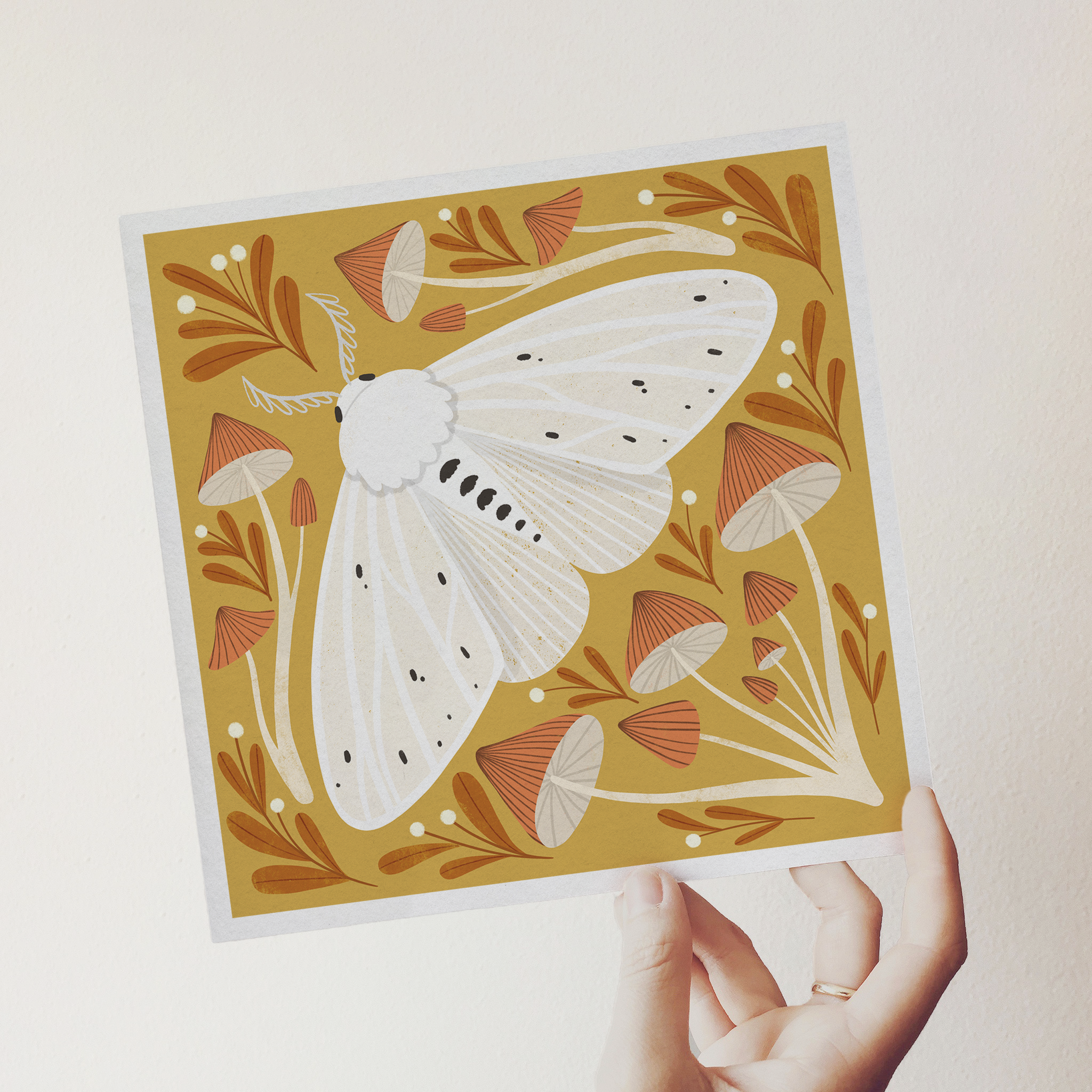 art print showing an agreeable tiger moth in black and white on a mustard yellow background, surrounded by red and white whimsical mushrooms. shown being held by a hand.