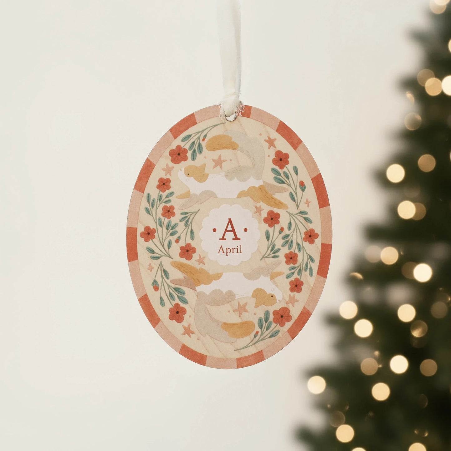 Illustrated wooden ornament with cats and flowers and the center monogram 'A' with 'April' text below, hanging against a blurred Christmas tree background. Ornament colors are primarily reds, greens, and white.