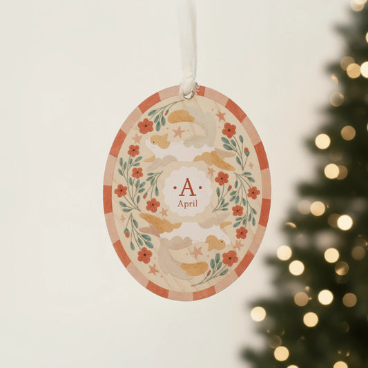 Illustrated wooden ornament with cats and flowers and the center monogram 'A' with 'April' text below, hanging against a blurred Christmas tree background. Ornament colors are primarily reds, greens, and white.