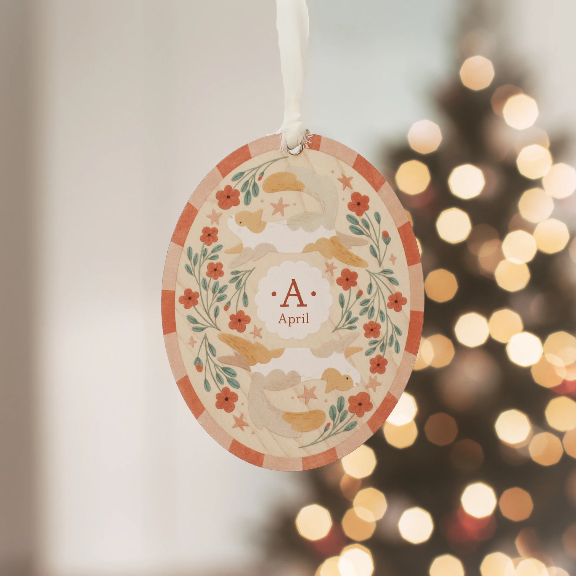 Illustrated wooden ornament with cats and flowers and the center monogram 'A' with 'April' text below, hanging against a blurred Christmas tree background. Ornament colors are primarily reds, greens, and whites.