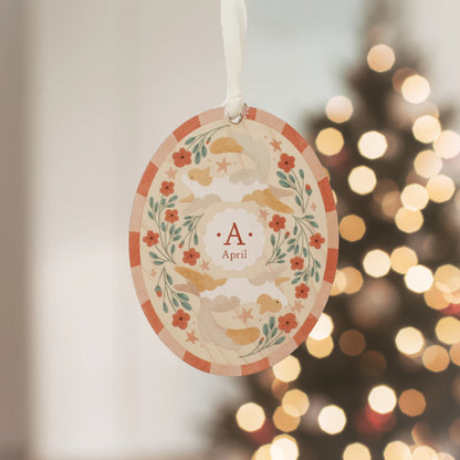 Illustrated wooden ornament with cats and flowers and the center monogram 'A' with 'April' text below, hanging against a blurred Christmas tree background. Ornament colors are primarily reds, greens, and whites.