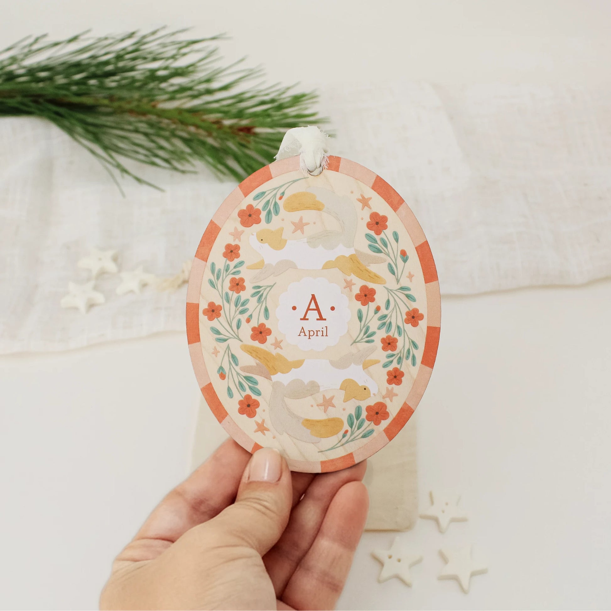 Illustrated wooden ornament with cats and flowers and the center monogram 'A' with 'April' text below, held by a hand against a light background. Ornament colors are primarily reds, greens, and whites.