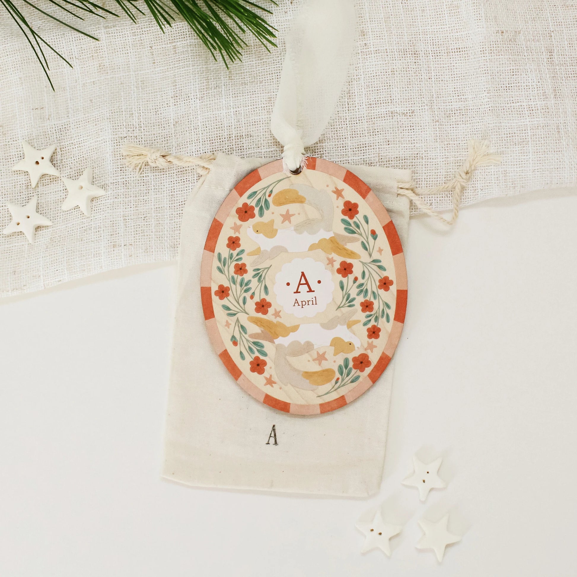 Illustrated wooden ornament with cats and flowers and the center monogram 'A' with 'April' text below, lying on a cotton bag stamped with the letter 'A' against a light background. Ornament colors are primarily reds, greens, and whites.