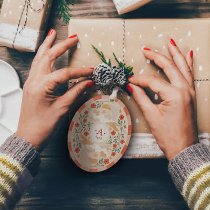 Person decorating a wrapped christmas gift with an Illustrated wooden ornament with cats and flowers and the center monogram 'A' with 'April' text below. Ornament colors are primarily reds, greens, and whites.