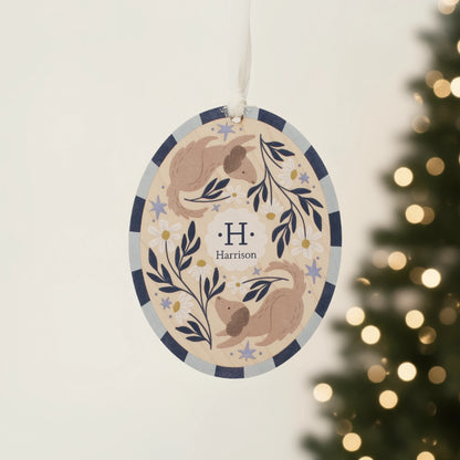 Illustrated wooden ornament with dogs and flowers and the center monogram 'H' with 'Harrison' text below, hanging against a blurred Christmas tree background. Ornament colors are primarily blues, yellow, and white. 