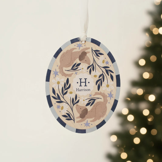 Illustrated wooden ornament with dogs and flowers and the center monogram 'H' with 'Harrison' text below, hanging against a blurred Christmas tree background. Ornament colors are primarily blues, yellow, and white. 