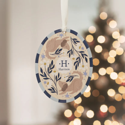 Illustrated wooden ornament with dogs and flowers and the center monogram 'H' with 'Harrison' text below, hanging against a blurred Christmas tree background. Ornament colors are primarily blues, yellow, and white.