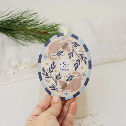 Illustrated wooden ornament with dogs and flowers and the center monogram 'S' with 'Sawyer' text below, held by a hand against a light background. Ornament colors are primarily blues, yellow, and white.