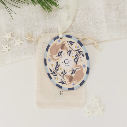 Illustrated wooden ornament with dogs and flowers and the center monogram 'G' with 'Greta' text below, lying on a cotton bag stamped with the letter 'G' against a light background. Ornament colors are primarily blues, yellow, and white.