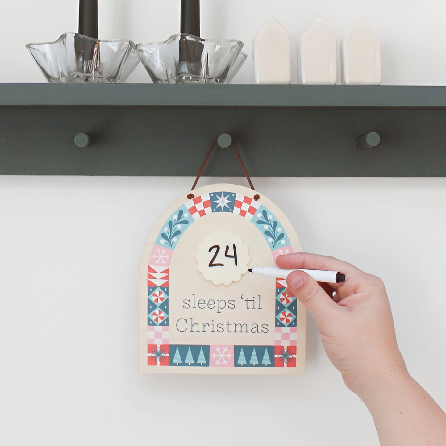 wood countdown sign with 'sleeps 'til Christmas' text and decorative patterns shown hanging on the wall while a hand writes 24 in the center