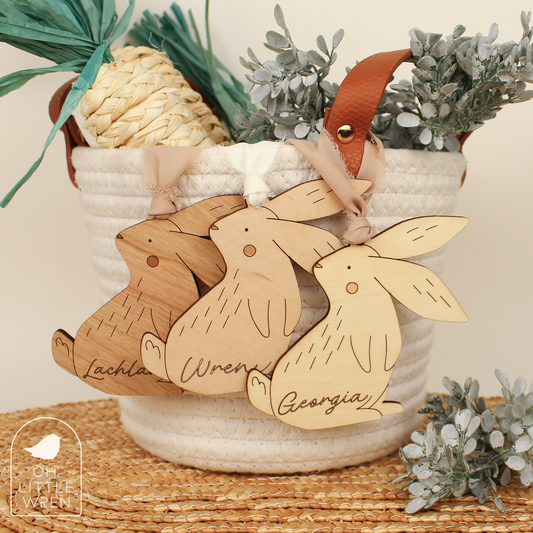 three wooden easter bunny tags, from left to right in cherry, maple, and birch wood tones, shown hanging from a white rope basket.