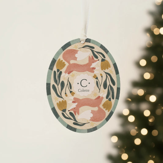 Illustrated wooden ornament with foxes and flowers and the center monogram 'C' with 'Colette' text below, hanging against a blurred Christmas tree background. Ornament colors are primarily greens, yellow, orange, and white. 