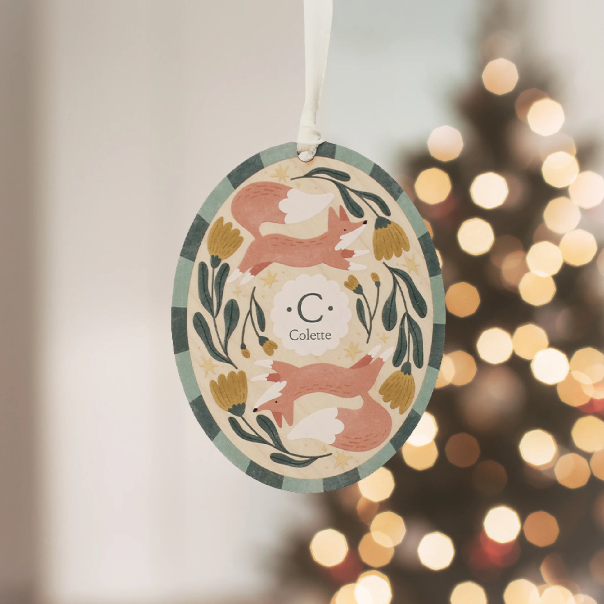 Illustrated wooden ornament with foxesand flowers and the center monogram 'C' with 'Colette' text below, hanging against a blurred Christmas tree background. Ornament colors are primarily greens, yellow, orange, and white. 