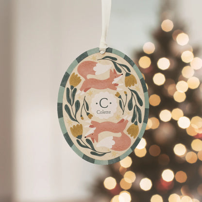 Illustrated wooden ornament with foxesand flowers and the center monogram 'C' with 'Colette' text below, hanging against a blurred Christmas tree background. Ornament colors are primarily greens, yellow, orange, and white. 