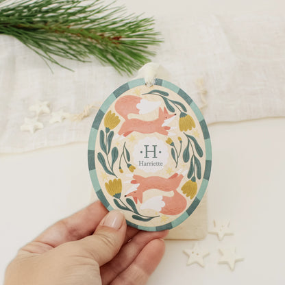 Illustrated wooden ornament with foxes and flowers and the center monogram 'H' with 'Harriette' text below, held by a hand against a light background. Ornament colors are primarily greens, yellow, orange, and white. 
