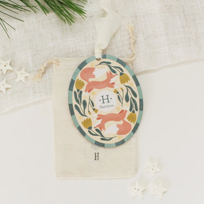 Illustrated wooden ornament with foxes and flowers and the center monogram 'H' with 'Harriette' text below, lying on a cotton bag stamped with the letter 'H' against a light background. Ornament colors are primarily greens, yellow, orange, and white. 