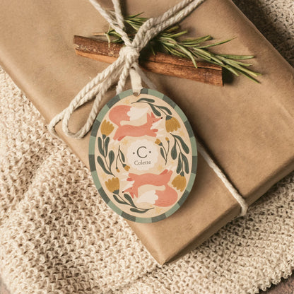 Illustrated wooden ornament with foxes and flowers and the center monogram 'C' with 'Colette' text below, being used to decorate a gift wrapped in brown kraft paper with a cinnamon stick and evergreen sprig, resting on a beige blanket. Ornament colors are primarily greens, yellow, orange, and white. 