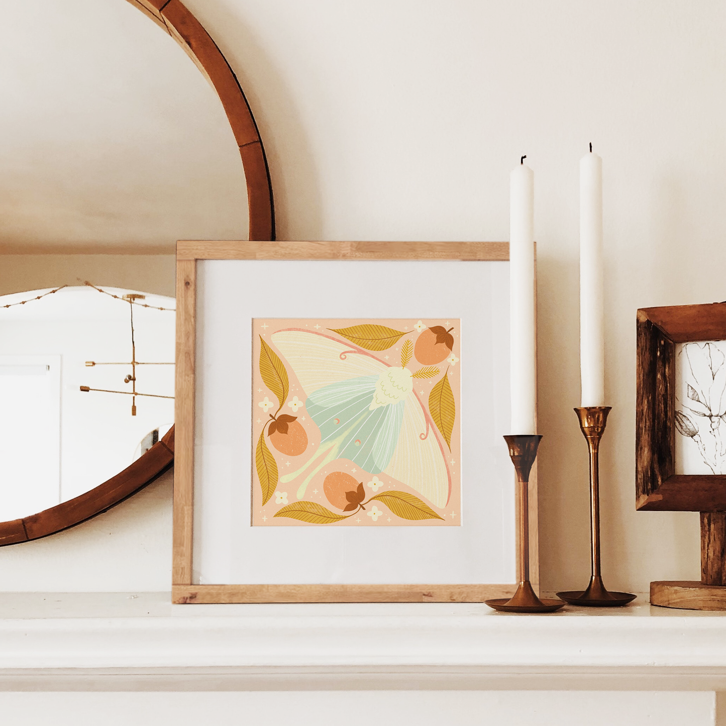 art print showing a luna moth in pale greens and pinks, surrounded by orange persimmons and mustard yellow leaves. shown displayed in a wood frame, resting on a mantle.