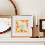 load image into gallery viewer, art print showing a luna moth in pale greens and pinks, surrounded by orange persimmons and mustard yellow leaves. shown displayed in a wood frame, resting on a mantle.

