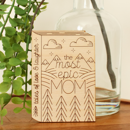 wooden book shaped card with forest and moutain motifs. cover reads 'the most epic mom'. spine reads 'tales of love & laughter'.