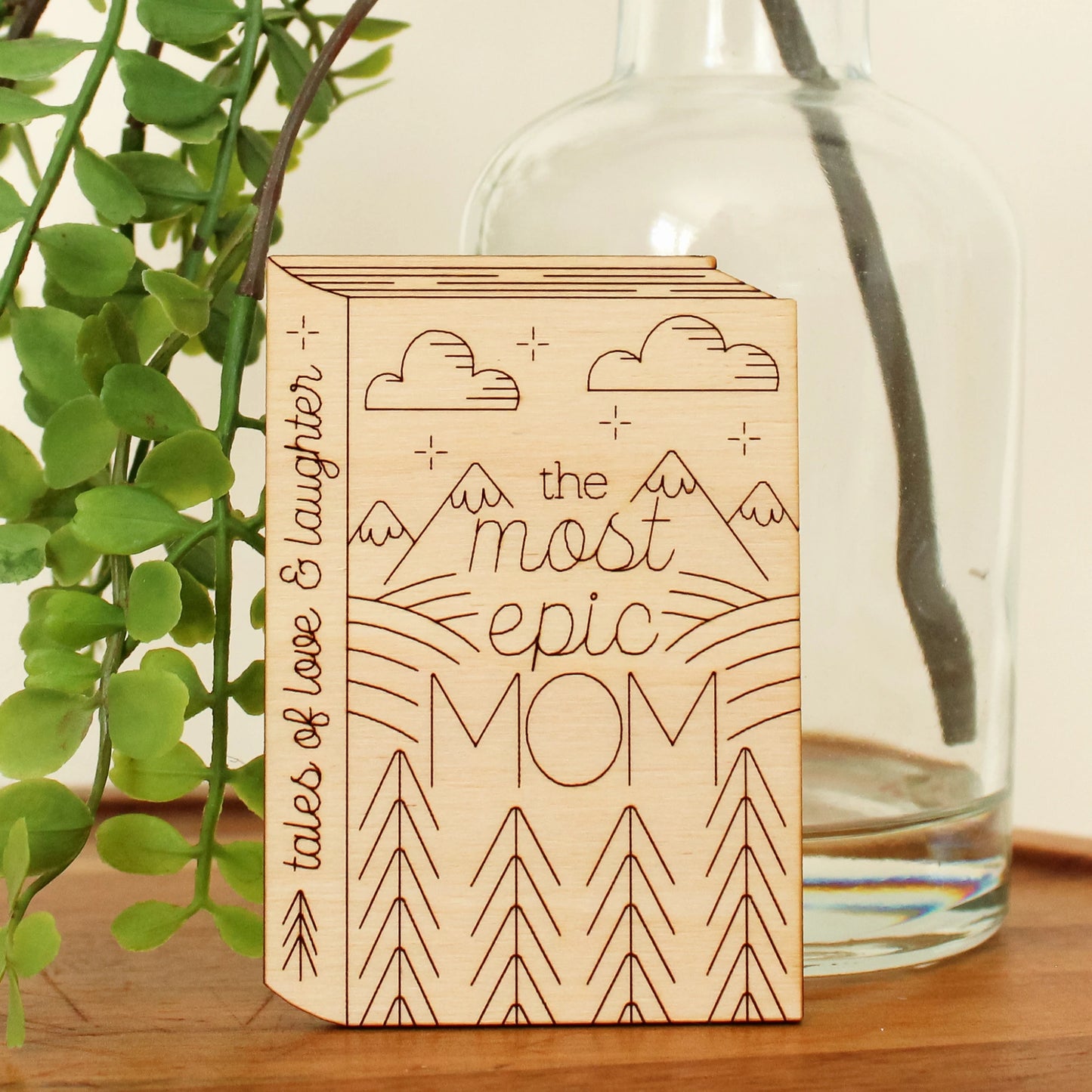 wooden book shaped card with forest and mountain motifs. cover reads 'the most epic mom'. spine reads 'tales of love & laughter'.