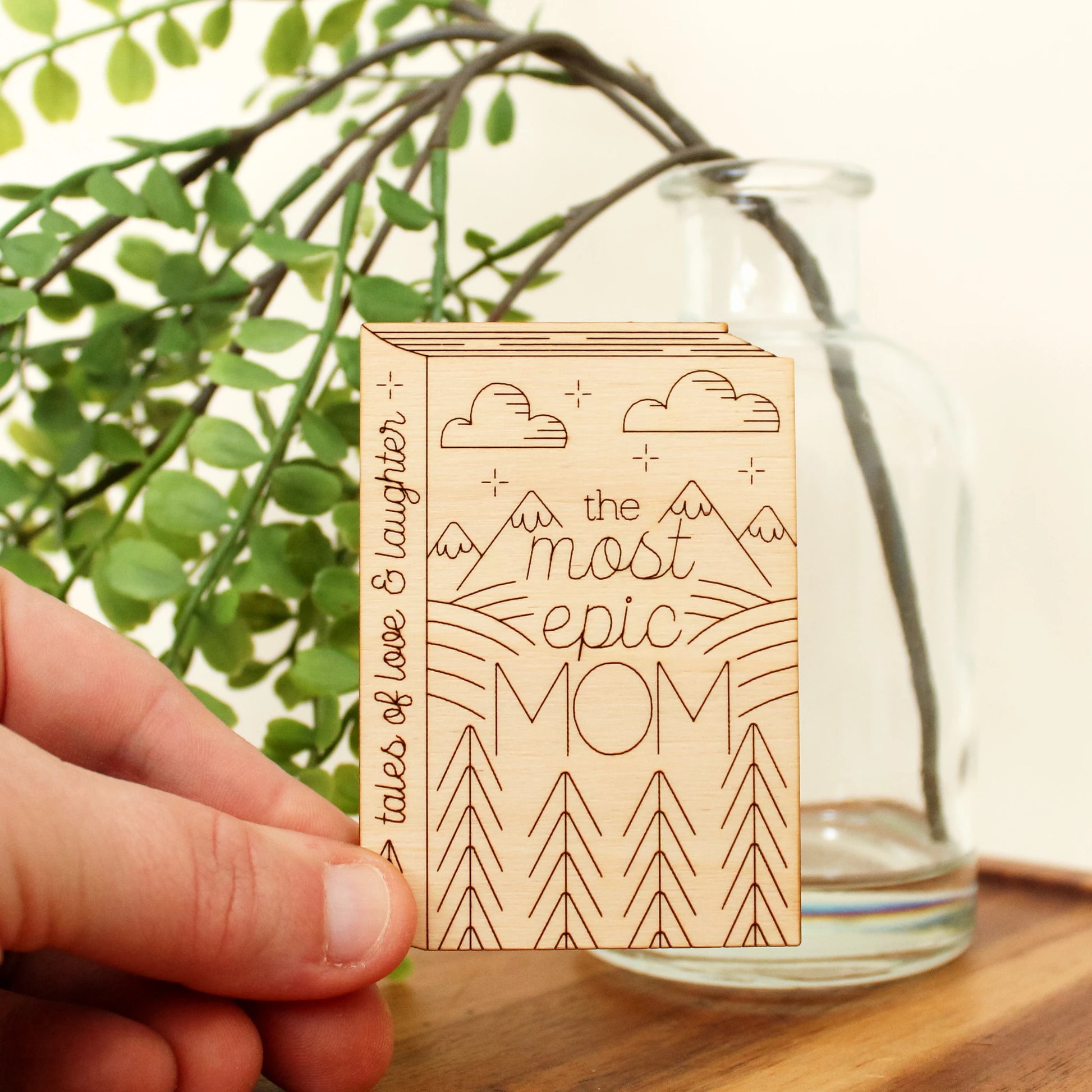 wooden book shaped card with forest and mountain motifs. cover reads 'the most epic mom'. spine reads 'tales of love & laughter'.