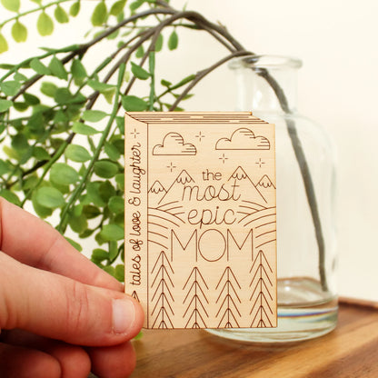 wooden book shaped card with forest and mountain motifs. cover reads 'the most epic mom'. spine reads 'tales of love & laughter'.