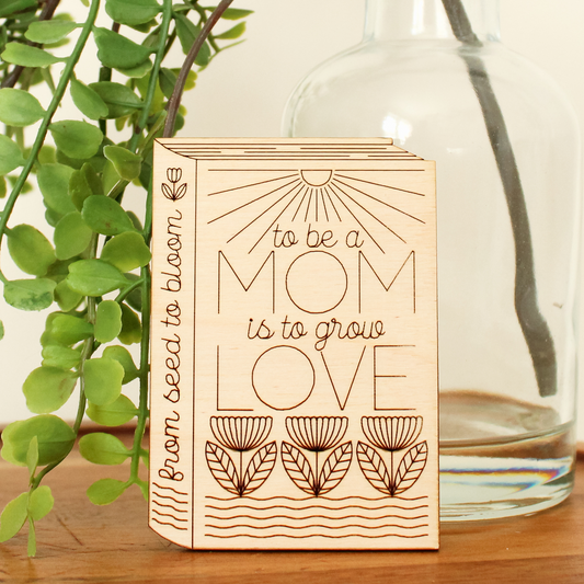wooden book shaped card with floral motifs. cover reads 'to be a mom is to grow love'. spine reads 'from seed to bloom'.