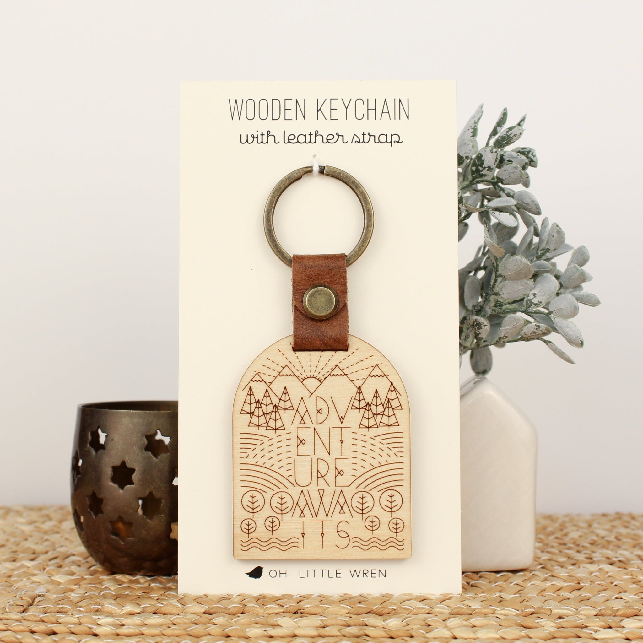 wooden keychain for the adventure lover. design has mountains, trees, fields and streams and the words 'adventure awaits'.  wooden keychain with leather strap and bronze colored hardware.