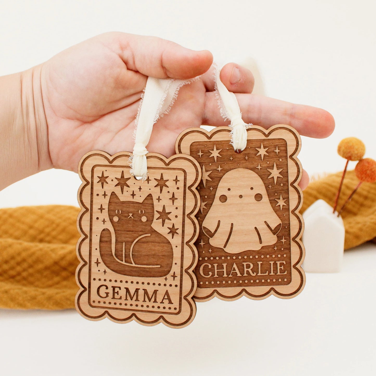two personalized halloween tags being held in front of an orange blanket. tag on the left has a black cat design and the name GEMMA and on the right is a ghost design with the name CHARLIE.