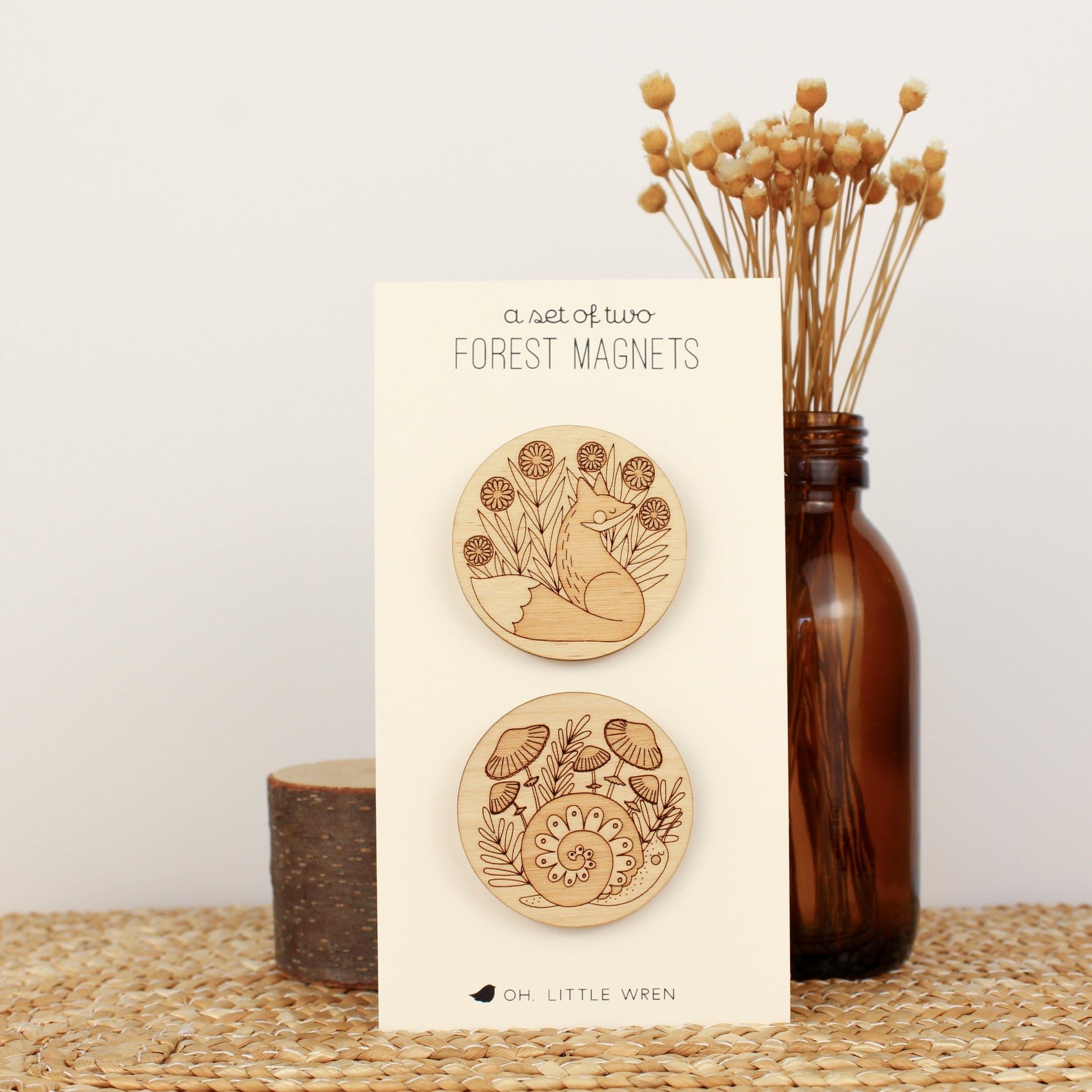 pair of circular wooden magnets, one with a fox surrounded by flowers and the other with a snail and mushrooms.
