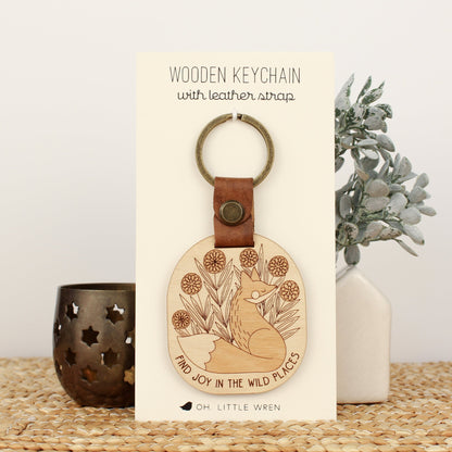 wooden keychain with fox design surrounded by flowers and the text 'find joy in the wild places.' with leather strap and bronze colored details.