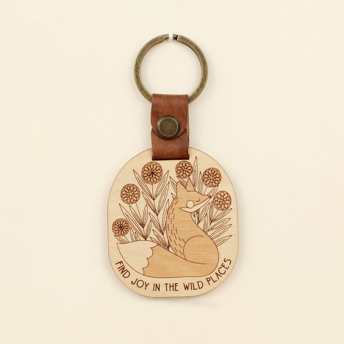 wooden keychain with fox design surrounded by flowers and the text 'find joy in the wild places.' with leather strap and bronze colored details.