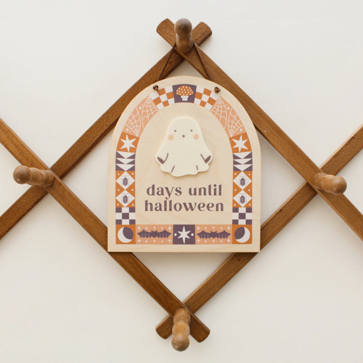 wooden countdown sign with text 'days until halloween' and a ghost-shaped spot for writing with a dry erase marker hangs on a wooden peg rack. design on sign features black and orange quilt motifs in an arch shape with halloween designs like bats, bugs, spider webs and more.