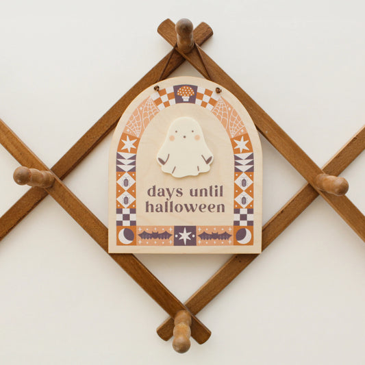 wooden countdown sign with text 'days until halloween' and a ghost-shaped spot for writing with a dry erase marker hangs on a wooden peg rack. design on sign features black and orange quilt motifs in an arch shape with halloween designs like bats, bugs, spider webs and more.