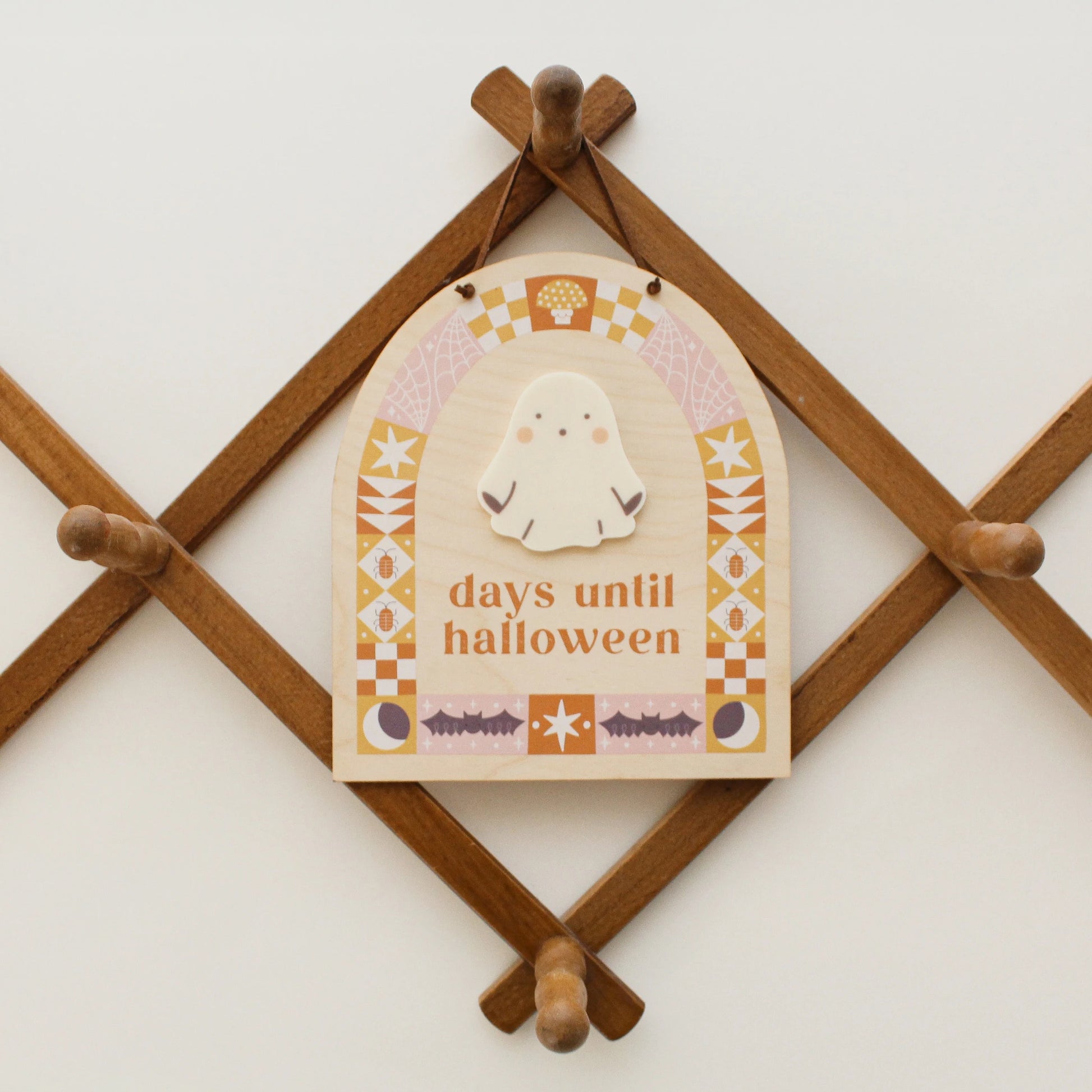 wooden countdown sign with text 'days until halloween' and a ghost-shaped spot for writing with a dry erase marker hangs on a wooden peg rack. design on sign features pink, orange and yellow quilt motifs in an arch shape with halloween designs like bats, bugs, spider webs and more.