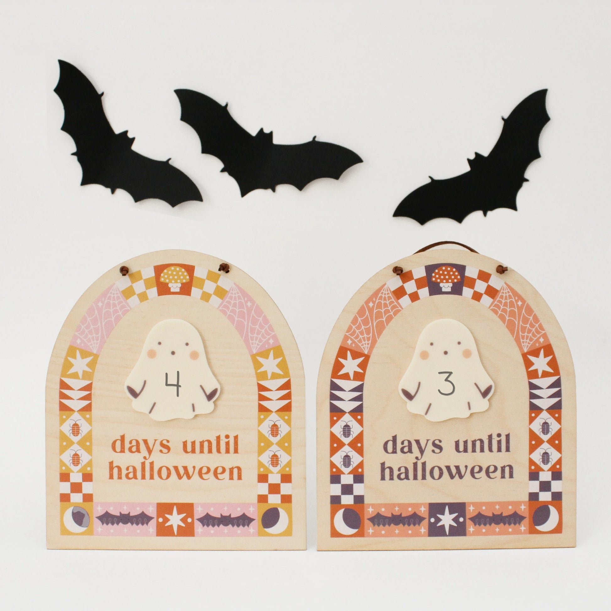 two wooden countdown signs with text 'days until halloween' and a ghost-shaped spot for writing with a dry erase marker are propped up on a white background. design on sign features quilt motifs in an arch shape with halloween designs like bats, bugs, spider webs and more. sign on the left is pink + orange and on right is black + orange.