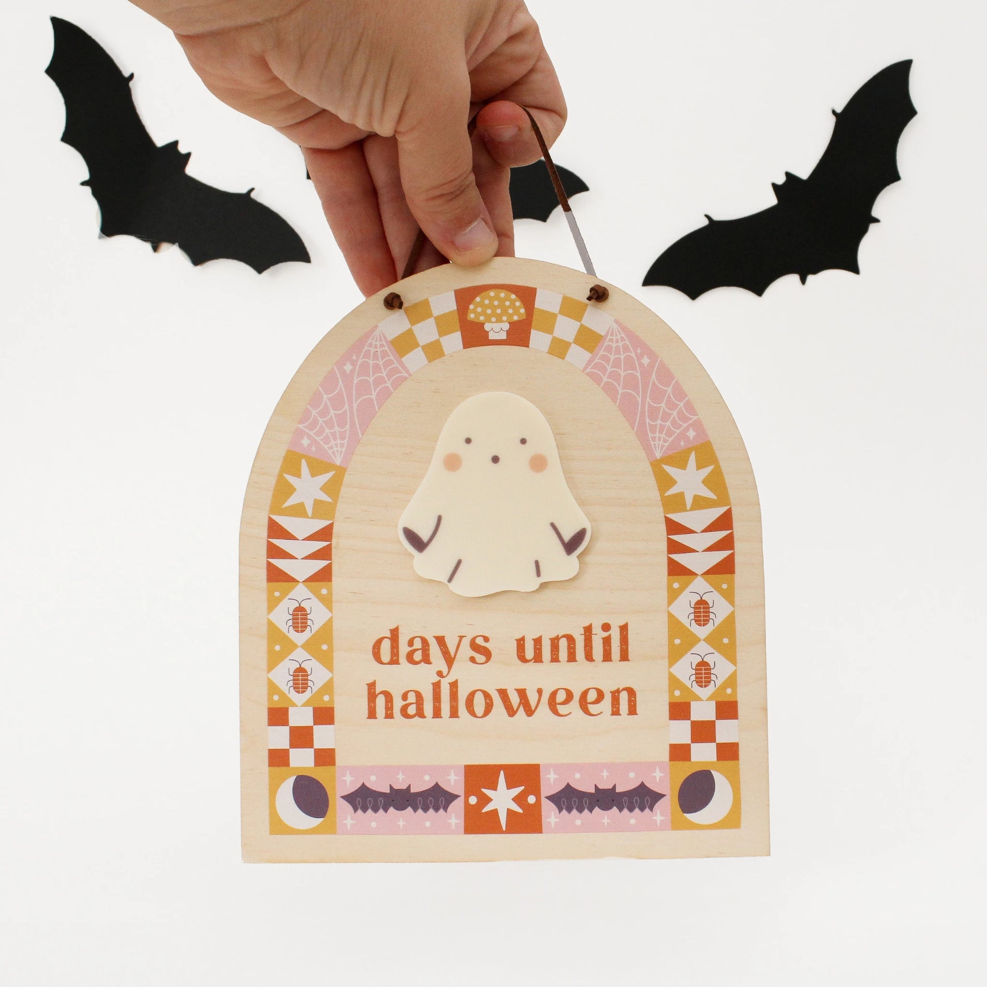 wooden countdown sign with text 'days until halloween' and a ghost-shaped spot for writing with a dry erase marker is held by a hand from the front to show scale. design on sign features pink, orange and yellow quilt motifs in an arch shape with halloween designs like bats, bugs, spider webs and more.