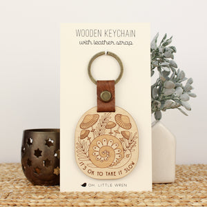 wooden keychain with snail design surrounded by mushrooms and the text 'it's ok to take it slow.' with leather strap and bronze colored details.