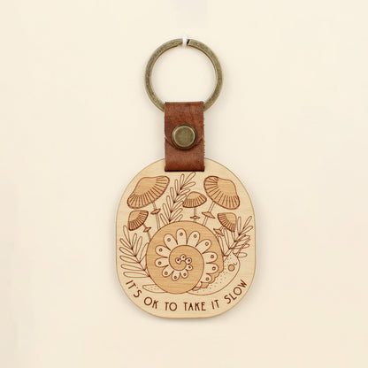 wooden keychain with snail design surrounded by mushrooms and the text 'it's ok to take it slow.' with leather strap and bronze colored details.