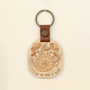 wooden keychain with snail design surrounded by mushrooms and the text 'it's ok to take it slow.' with leather strap and bronze colored details.
